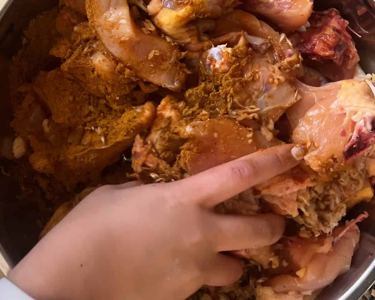 A hand mixes curry powder into raw chicken in a metal bowl