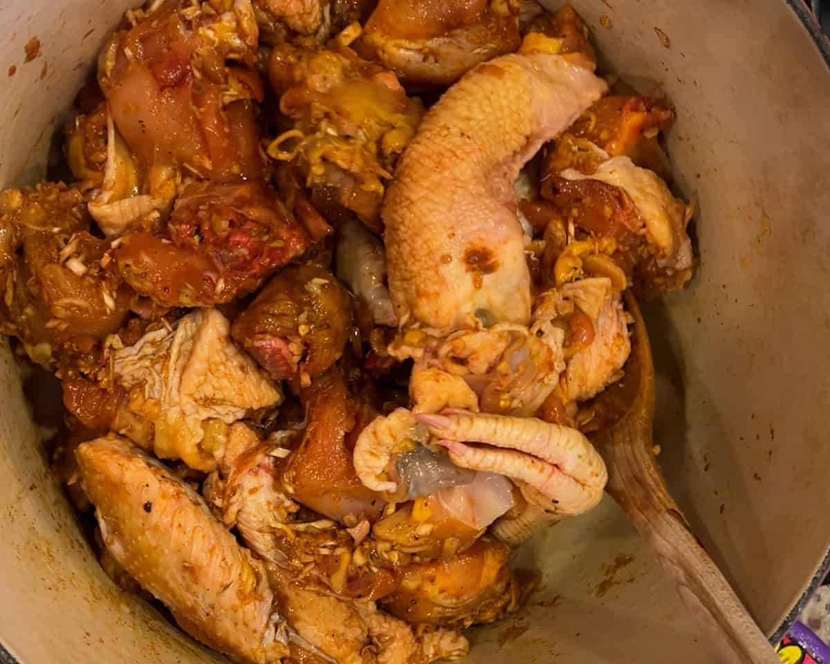 A wooden spoon mixes marinated chicken in a dutch oven