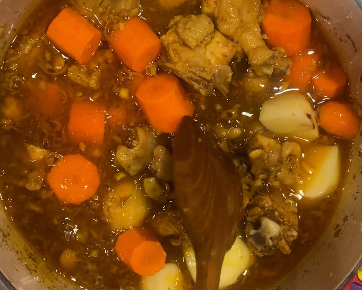 A wooden spoon stirs potatoes and carrots into a pot of chicken curry