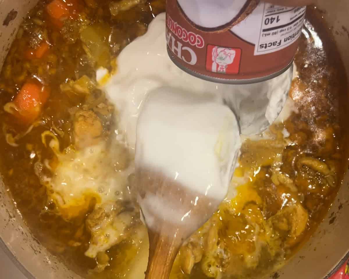A hand pours coconut milk into a pot of chicken curry