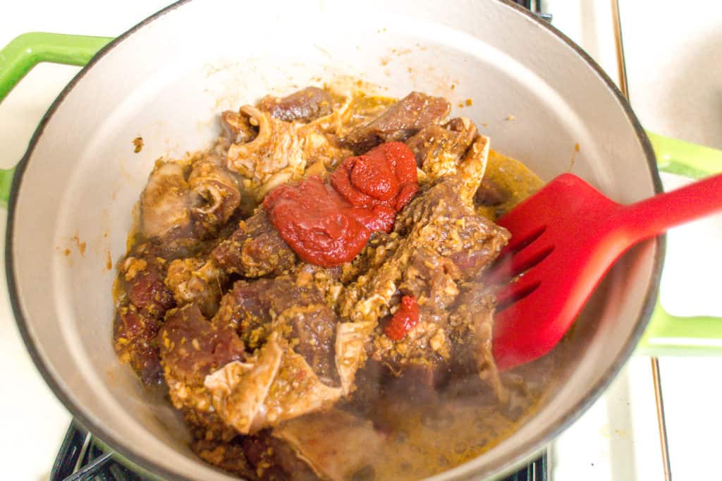 green dutch oven with beef cubes and tomato paste. A red spatula is pushing the meat around