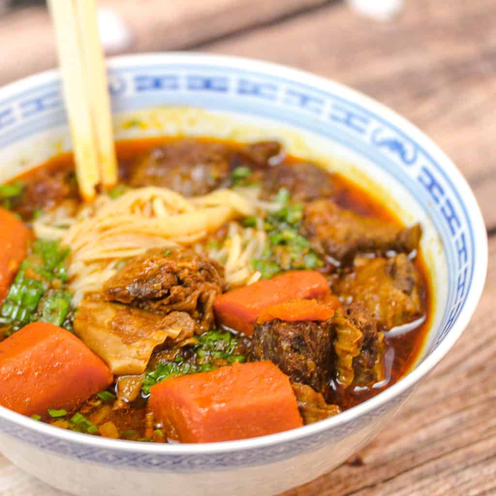 blue and white bowl filled with noodles, stew with beef and carrots. A pair of chopsticks is sticking into the noodles