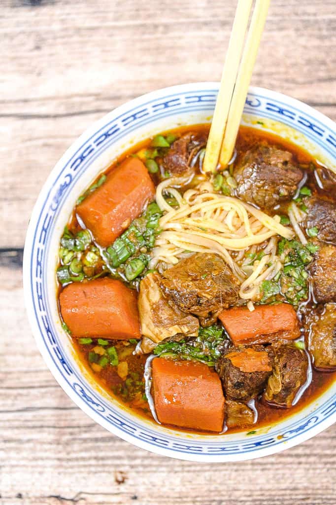 blue and white bowl filled with noodles, stew with beef and carrots. A pair of chopsticks is sticking into the noodles