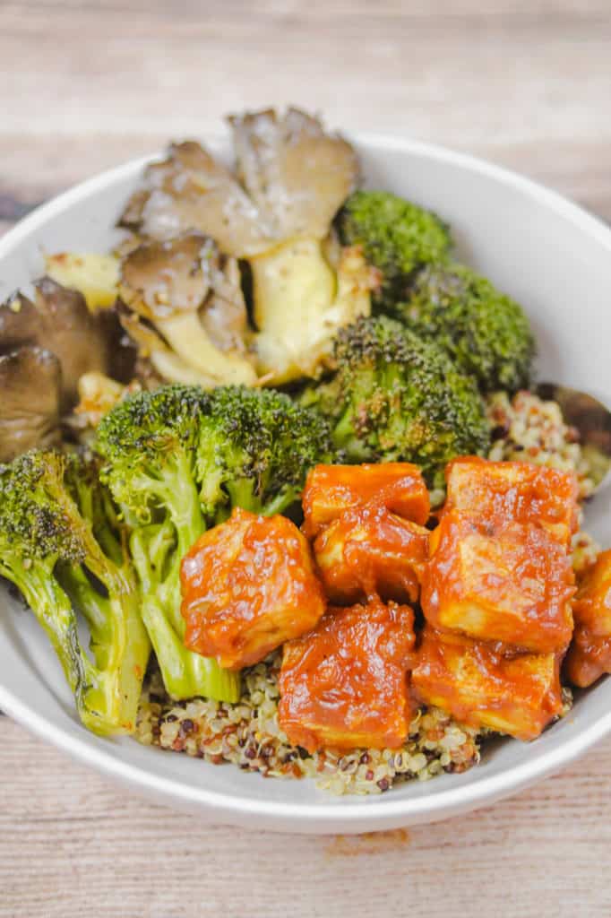 White bowl with tri-color quinoa, tofu cubes coated in peanut sauce, roasted broccoli and oyster mushrooms