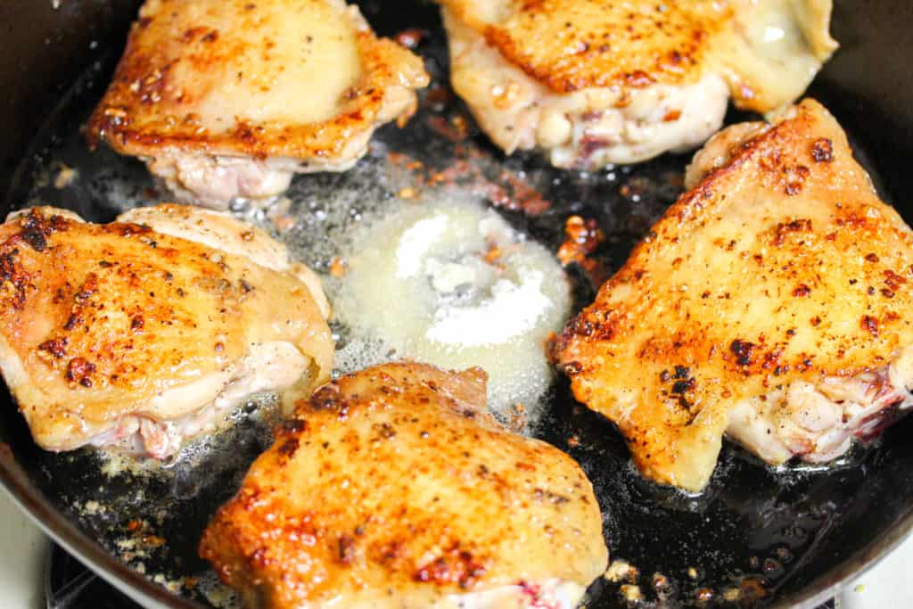 Crispy chicken thighs for Gà Roti cooking skin side up in a cast iron skillet. In the middle of the skillet is a mound of granulated sugar. 