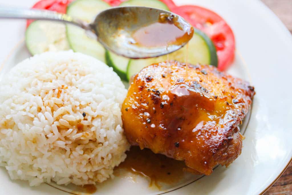 A Gà Roti plate: A white plate with one glazed chicken thigh, a round mound of rice, and cucumbers and tomatoes