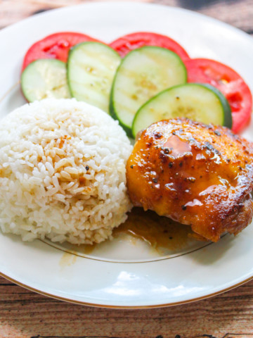 White plate with gold trim. On the plate is a chicken thigh with brown glaze, a round mound of rice, slices of tomato with slices of cucumber layered on top of the tomato.