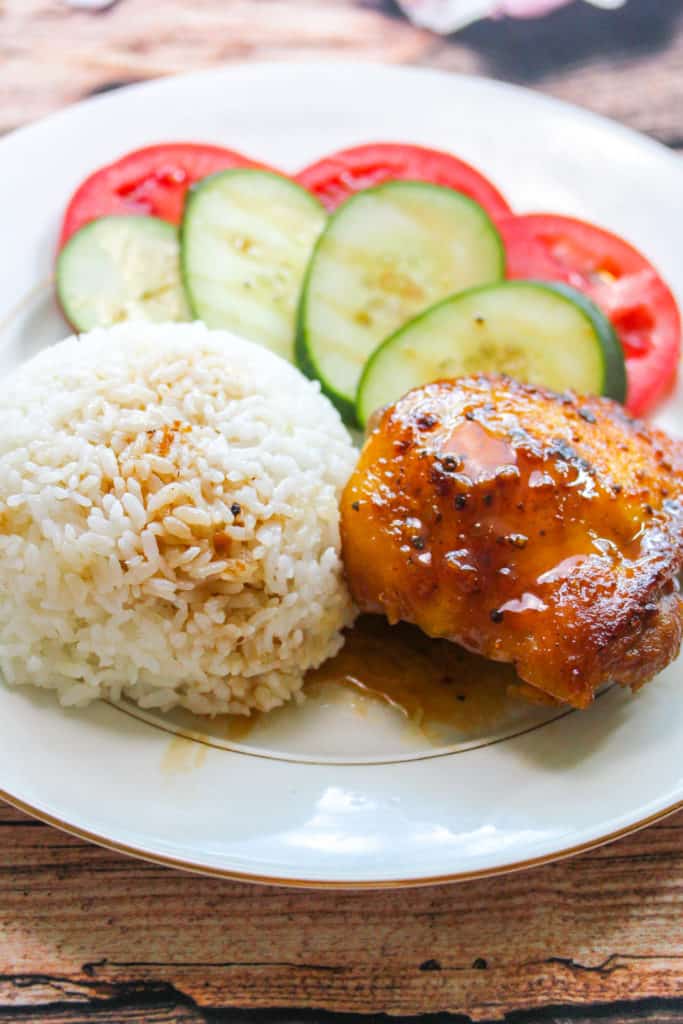 White plate with gold trim. On the plate is a chicken thigh (Gà Roti) with brown glaze, a round mound of rice, slices of tomato with slices of cucumber layered on top of the tomato. 