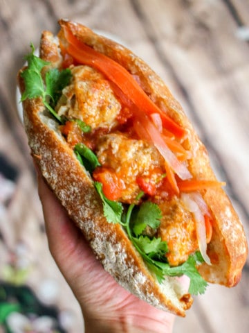 A hand holding a baguette sandwich with three meatballs, cilantro and pickled carrots and daikon radish