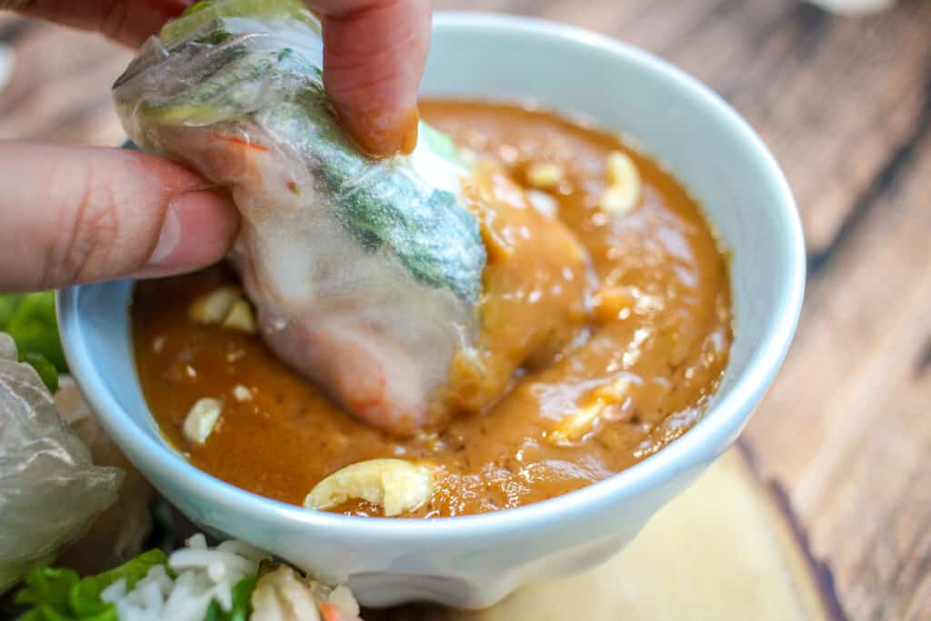A spring roll being dipped into a bowl of peanut sauce