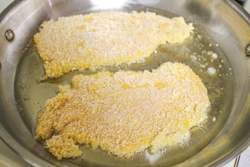 Two breaded chicken cutlets cooking in a stainless steel pan