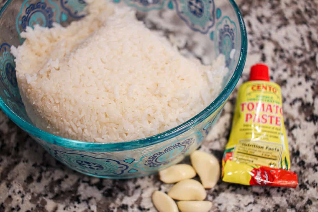 A bowl of rice next to a tube of tomato paste and garlic cloves