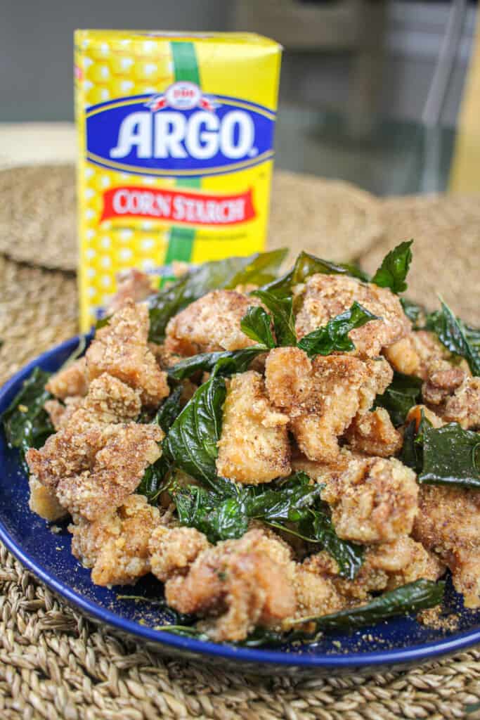 a straight on shot of a plate of taiwanese popcorn chicken with fried thai basil in front of a box of Argo corn starch