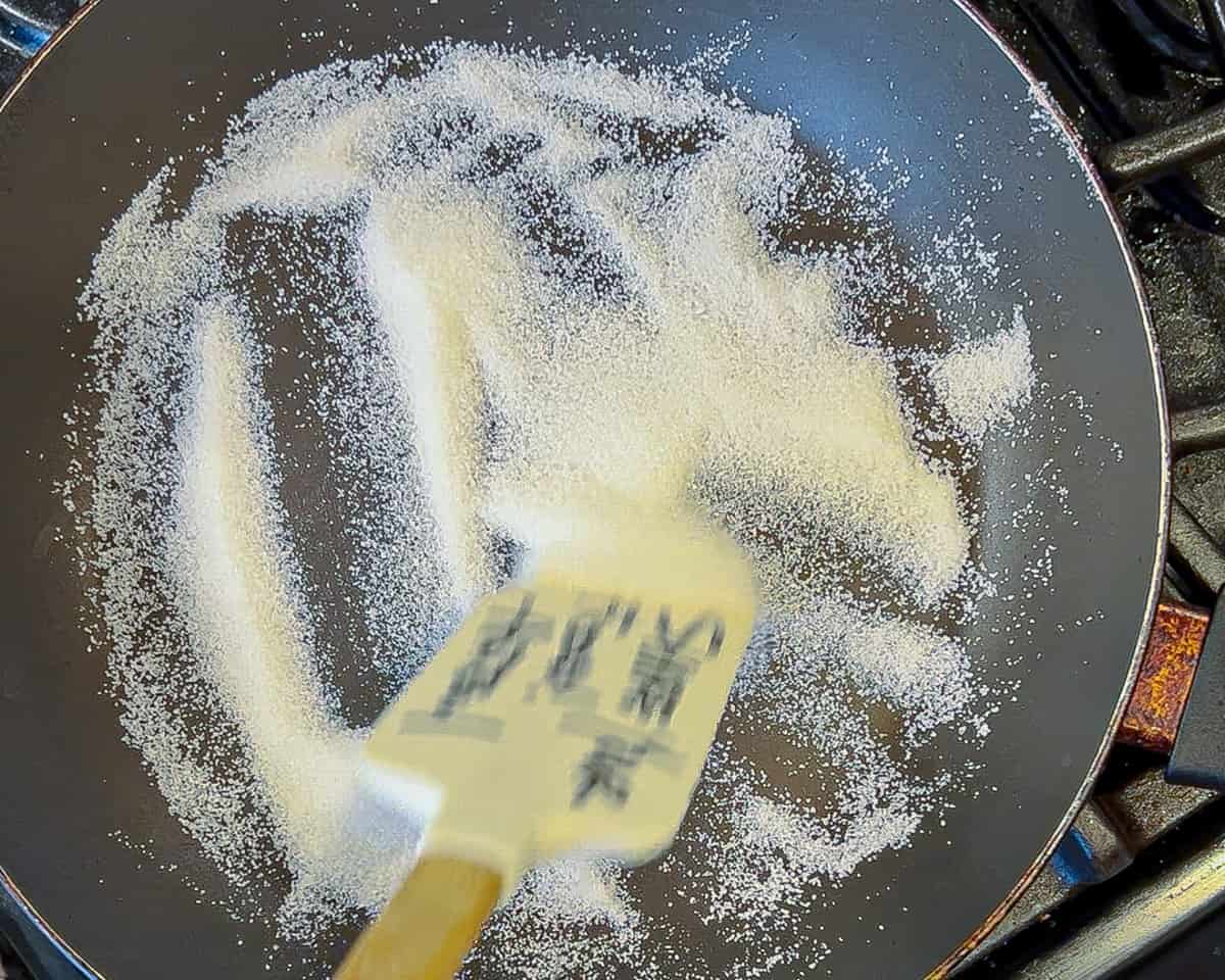 A wooden spatula stirs white sugar in a nonstick pan