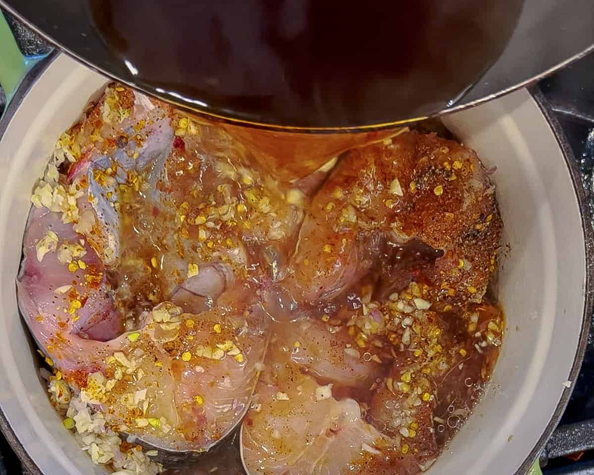 Brown liquid is poured over a dutch oven filled with raw marinated catfish steaks