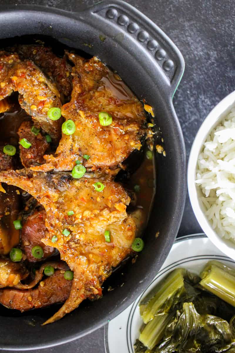 A closeup of a casserole dish full of Vietnamese Braised Catfish (Cá Kho Tộ) next to a plate of mustard greens and rice