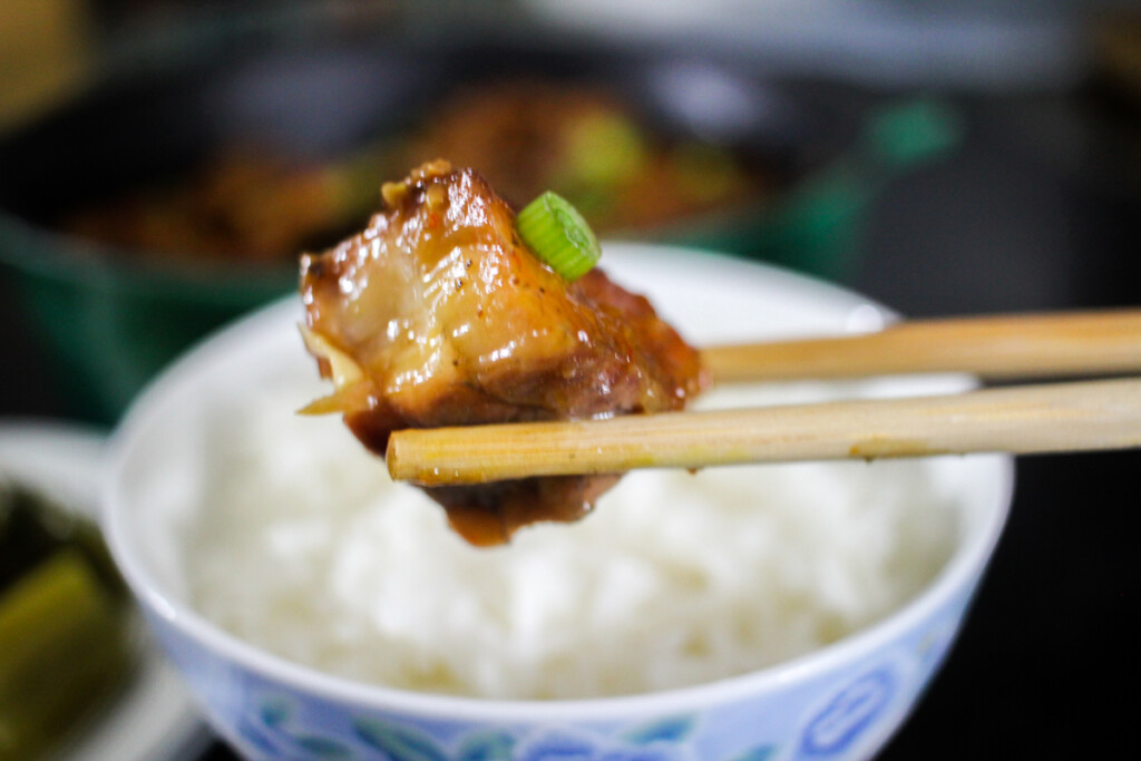 Chopsticks hold up a bite sized morsel of braised catfish in front of a small bowl of rice