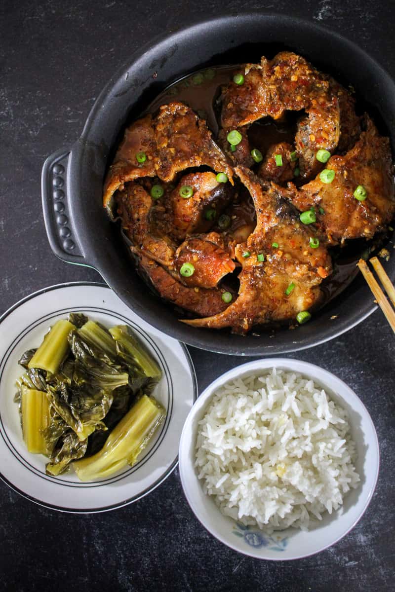 A bird's eye view of a round casserole dish filled with Vietnamese Braised Catfish (Cá Kho Tộ) next to a small plate of pickled mustard greens and a bowl of rice.