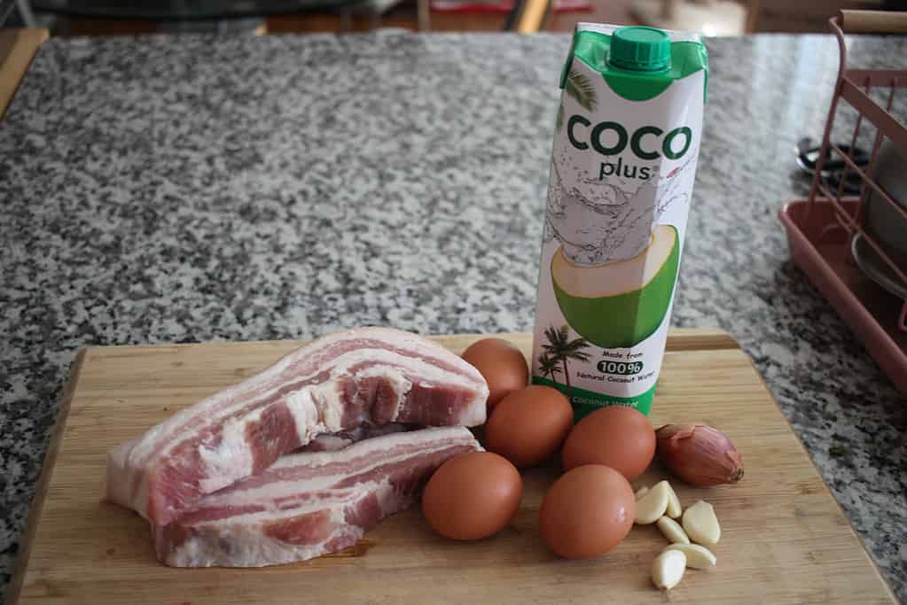 raw pork belly, eggs, garlic, shallot and a bottle of coconut water on top of a wooden cutting board.