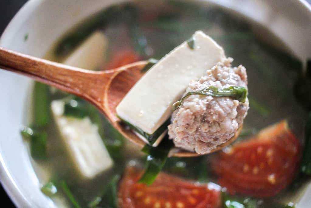 A close up of a wooden spoon with a piece of tofu and a pork meatball over a bowl of Vietnamese Tofu Soup with Tomato and Chives (Canh hẹ Đậu Hũ Cà Chua)