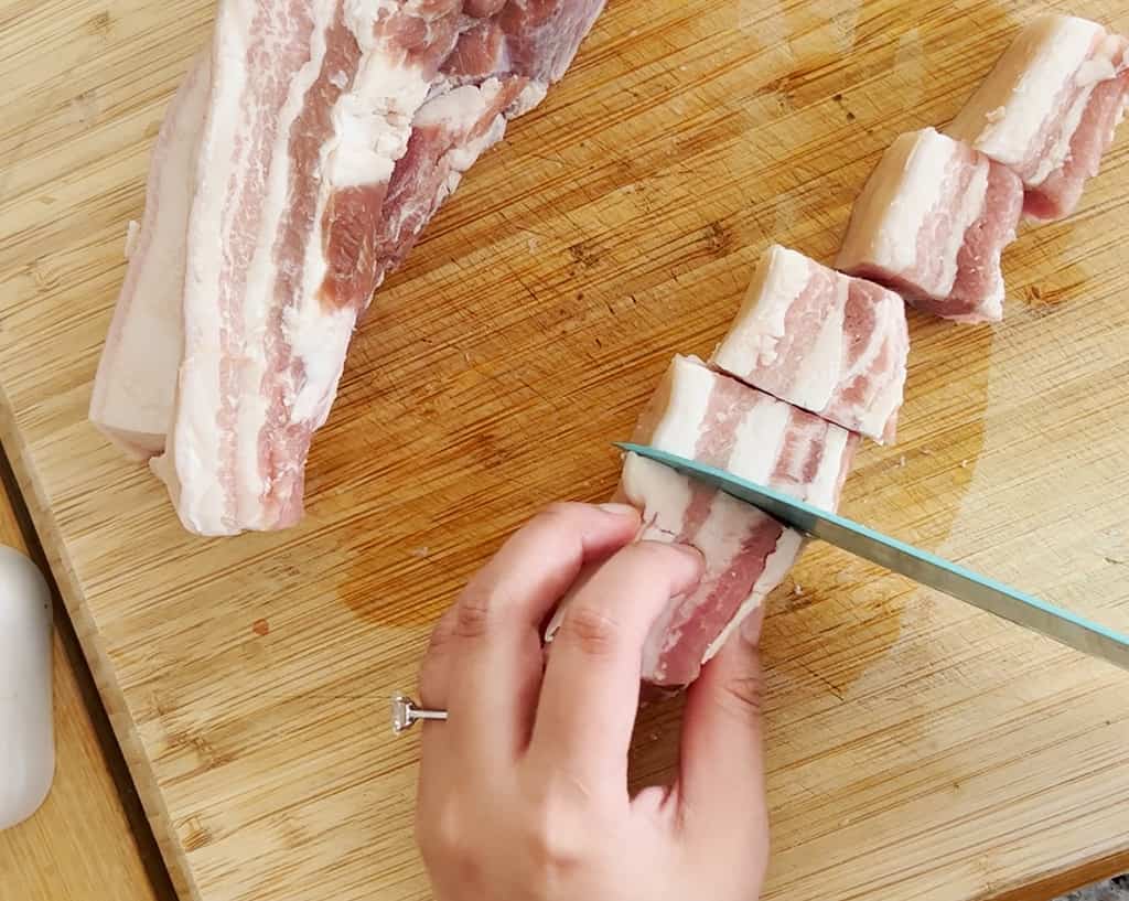 A hand chops raw pork belly into chunks