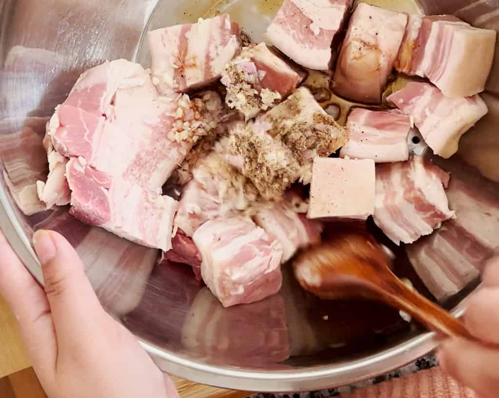 A wooden spoon stirs raw chunks of pork belly in a metal bowl