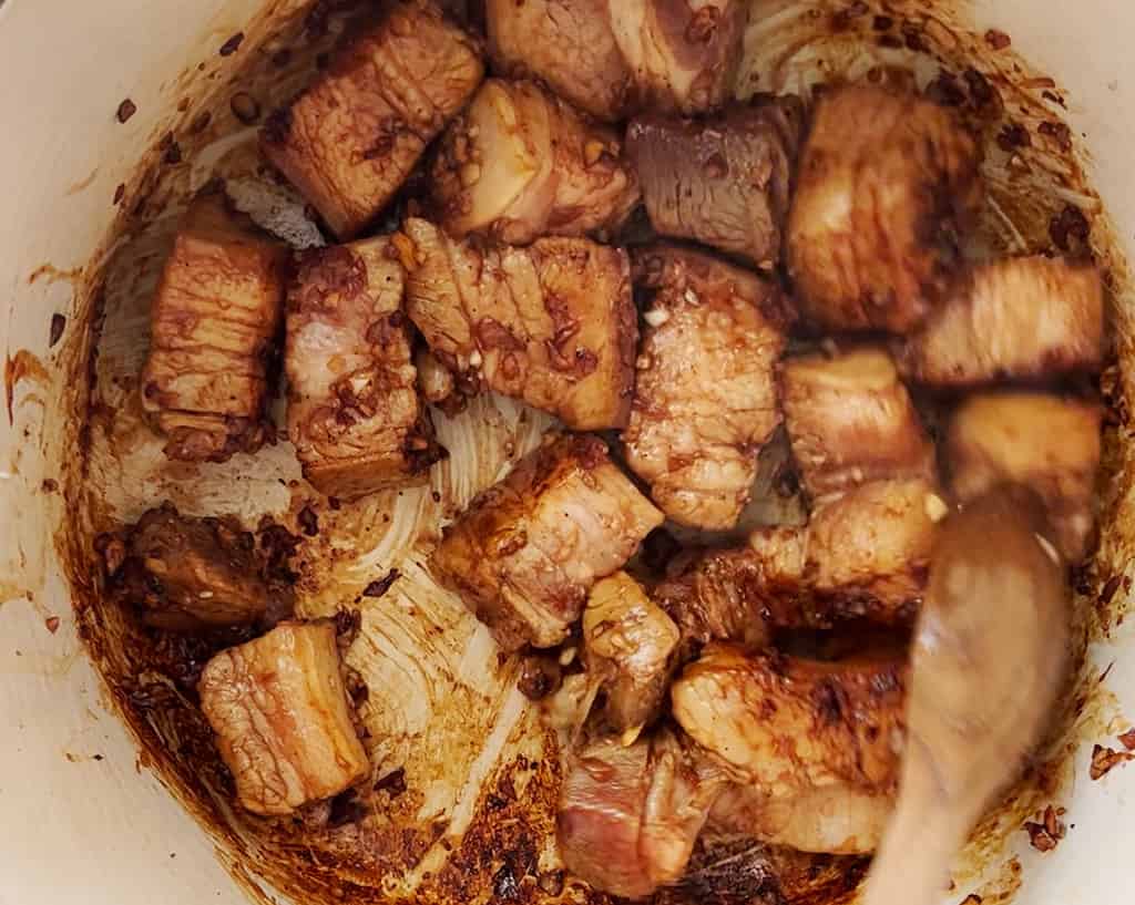 A wooden spoon stirs chunks of pork belly, coating them in caramelized sugar