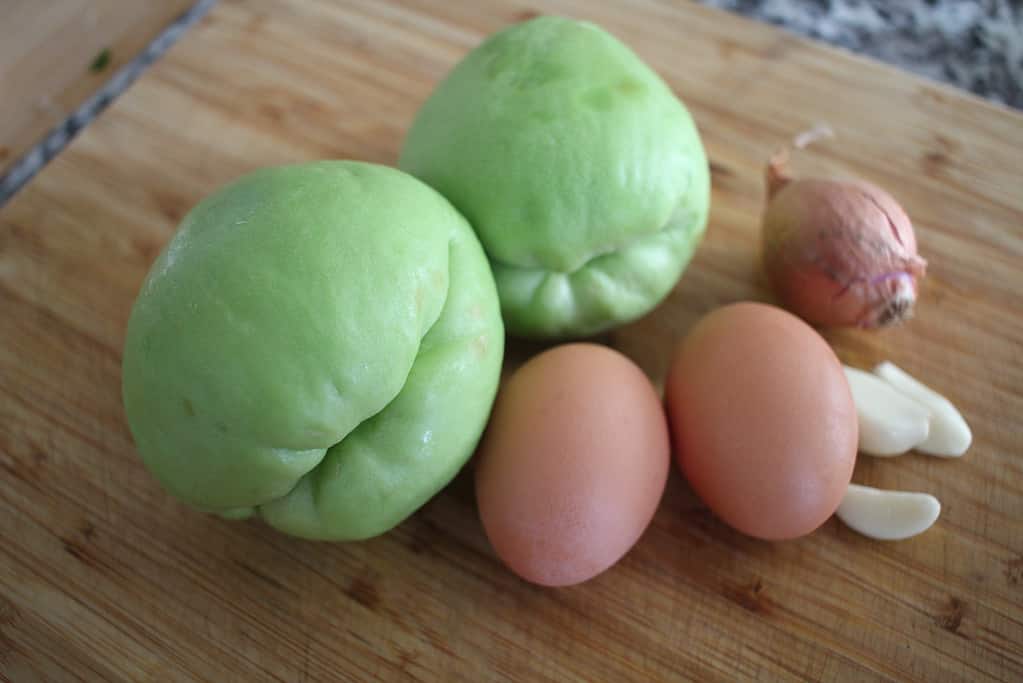 two chayote squash, two egg, a shallot and some garlic cloves on a wooden cutting board