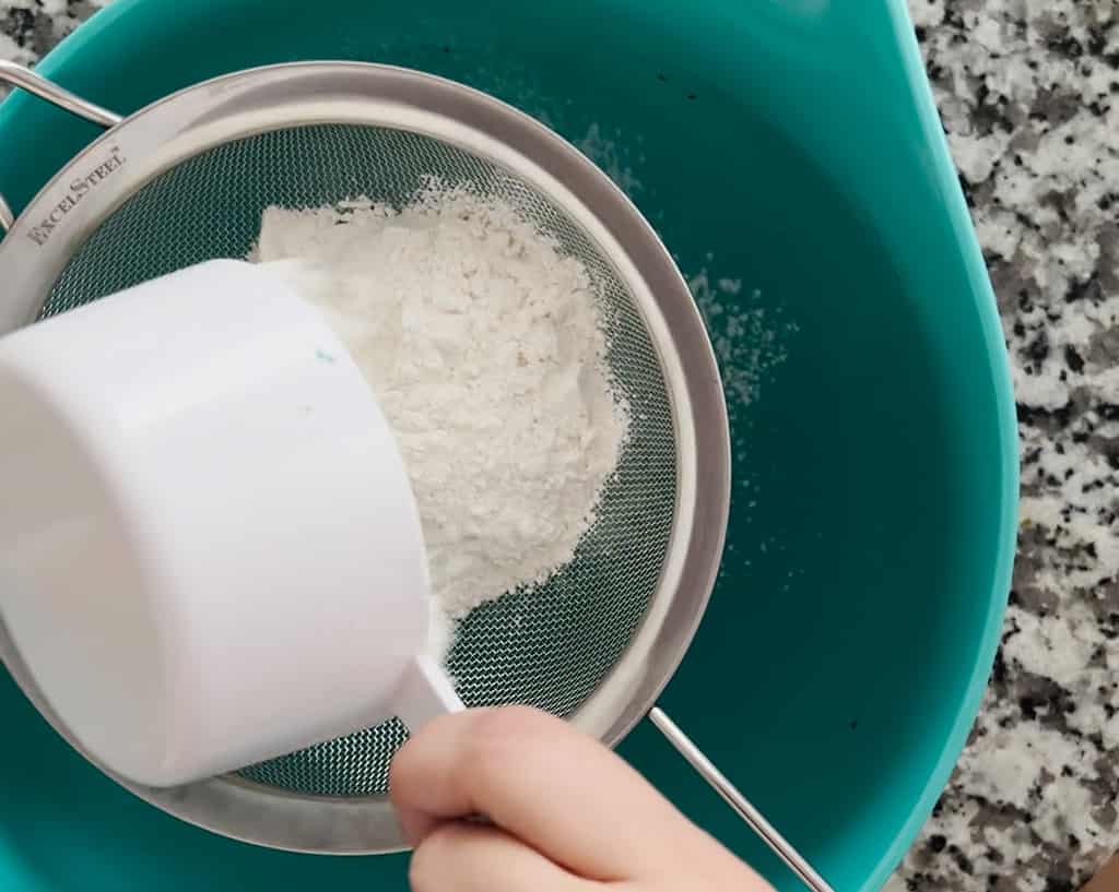 A bird's eye view of a person pouring flour into a sieve sitting on top of a blue bowl.