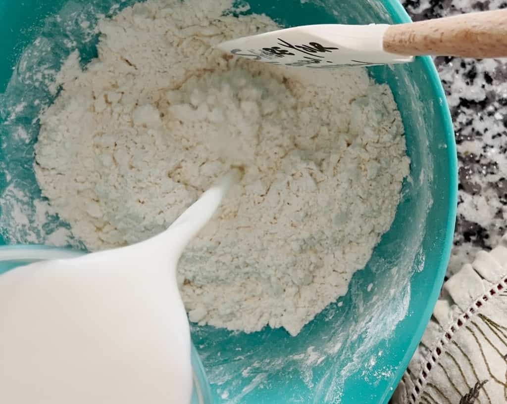 A bird's eye view of a person pouring buttermilk into a blue bowl of butter mixed with flour.