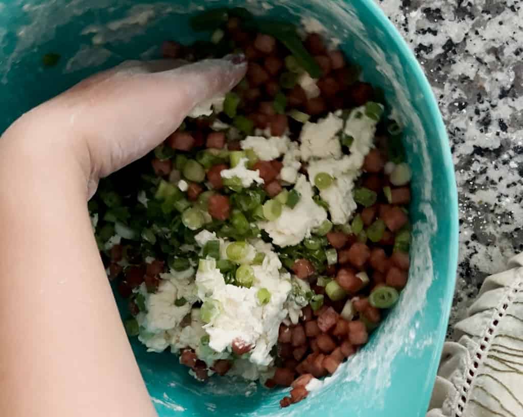 A bird's eye view of a person mixing chopped scallion and diced crispy spam into a dough.