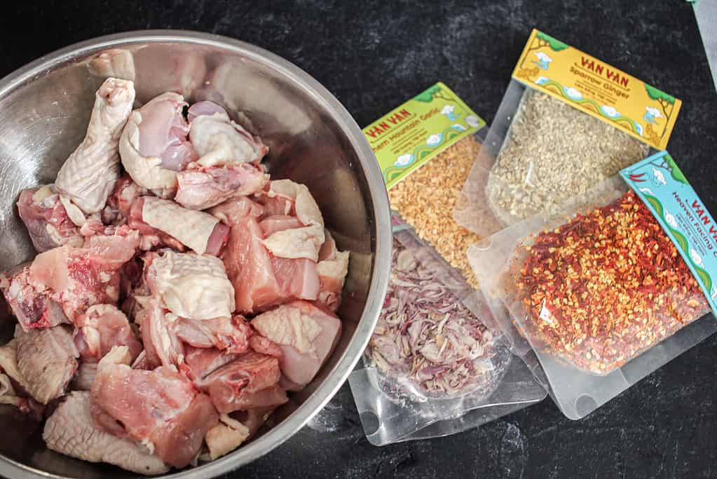 A metal bowl full of chopped raw chicken sits next to packets of dried chili, shallot, garlic and ginger.