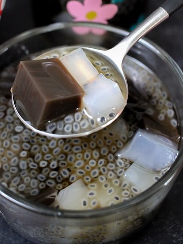A square cropped, close up shot of a metal spoon holds a chunk of grass jelly, two chunks of nata de coco and some basil seeds in sugar syrup. It is held above a clear bowl with more grass jelly, nata de coco and basil seeds.