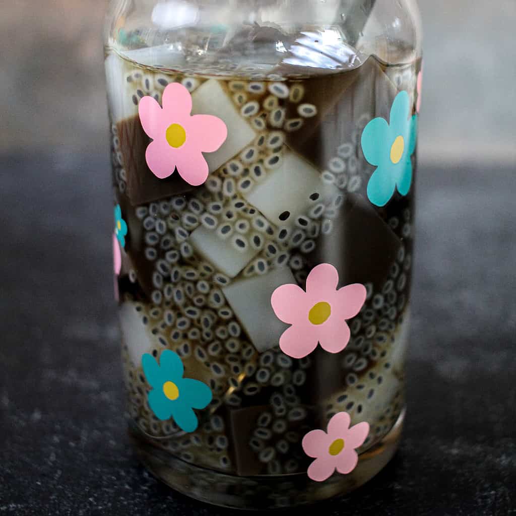 A tall clear glass with blue and pink flowers holds grass jelly with basil seeds and nata de coco suspended in sugar water. It is placed on a black background.