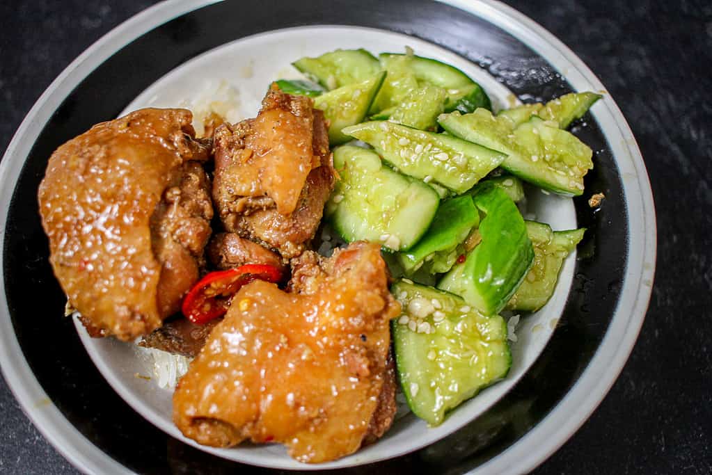 A white bowl with a black trim is filled with rice, cucumber salad and ginger-braised chicken