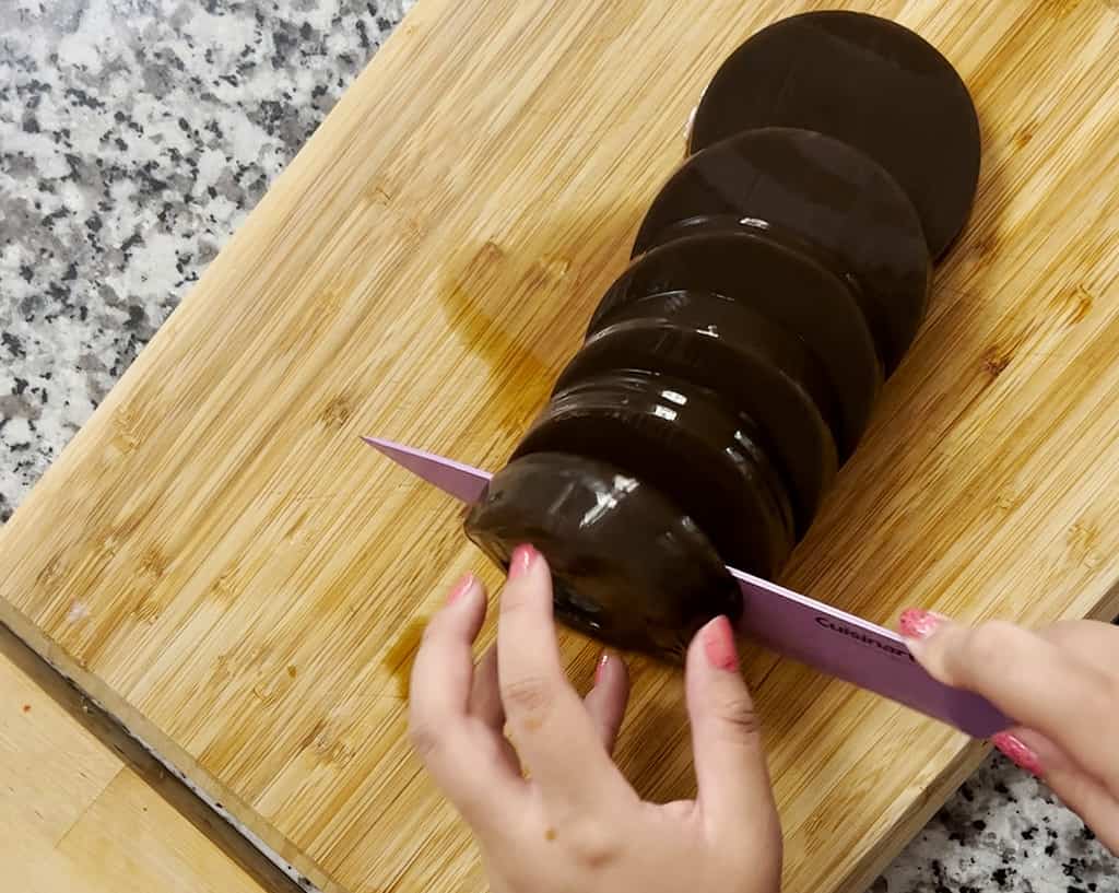Two hands holding a purple knife cut a cylinder of grass jelly into even slices.