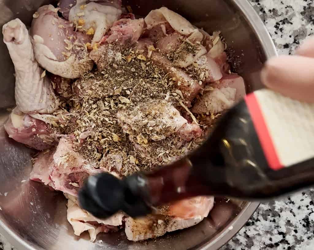 A hand pours fish sauce from a glass bottle into a metal bowl filled with chopped chicken, garlic, ginger, shallot, salt and pepper.