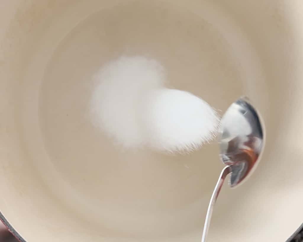 A metal spoon pours granulated sugar into a dutch oven