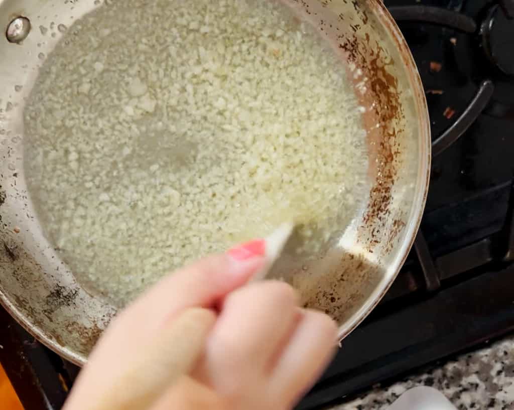 A hand stirs minced garlic sizzling in oil in a stainless steel pan