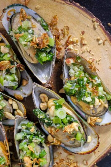 A square crop of Five Grilled Scallion Oil Mussels (Chem Chép Nướng Mỡ Hành) arranged on a board that looks like a slice of a tree trunk. Fried shallots are strewn on top of the board.