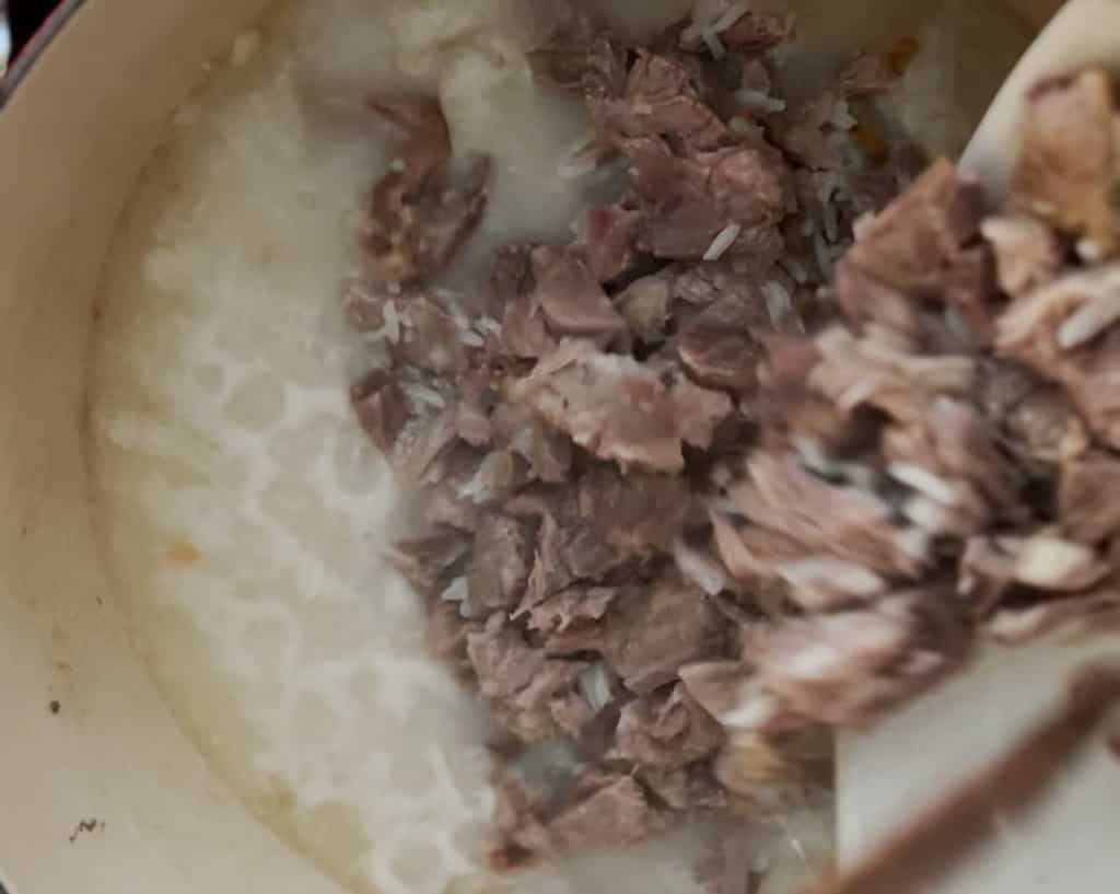 A bowl dumps shredded pork bone meat into a pot of congee
