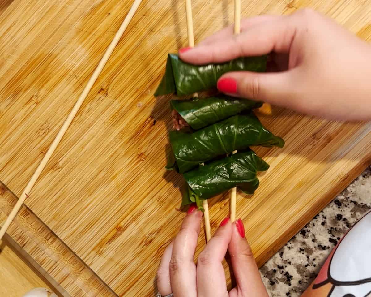 Two hands attach betel leaf wrapped beef pieces onto two skewers.