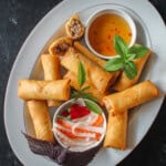 Plate of golden, crispy Vietnamese chả giò (egg rolls) served with fresh herbs, pickled carrots and daikon, and a small bowl of nước chấm dipping sauce.