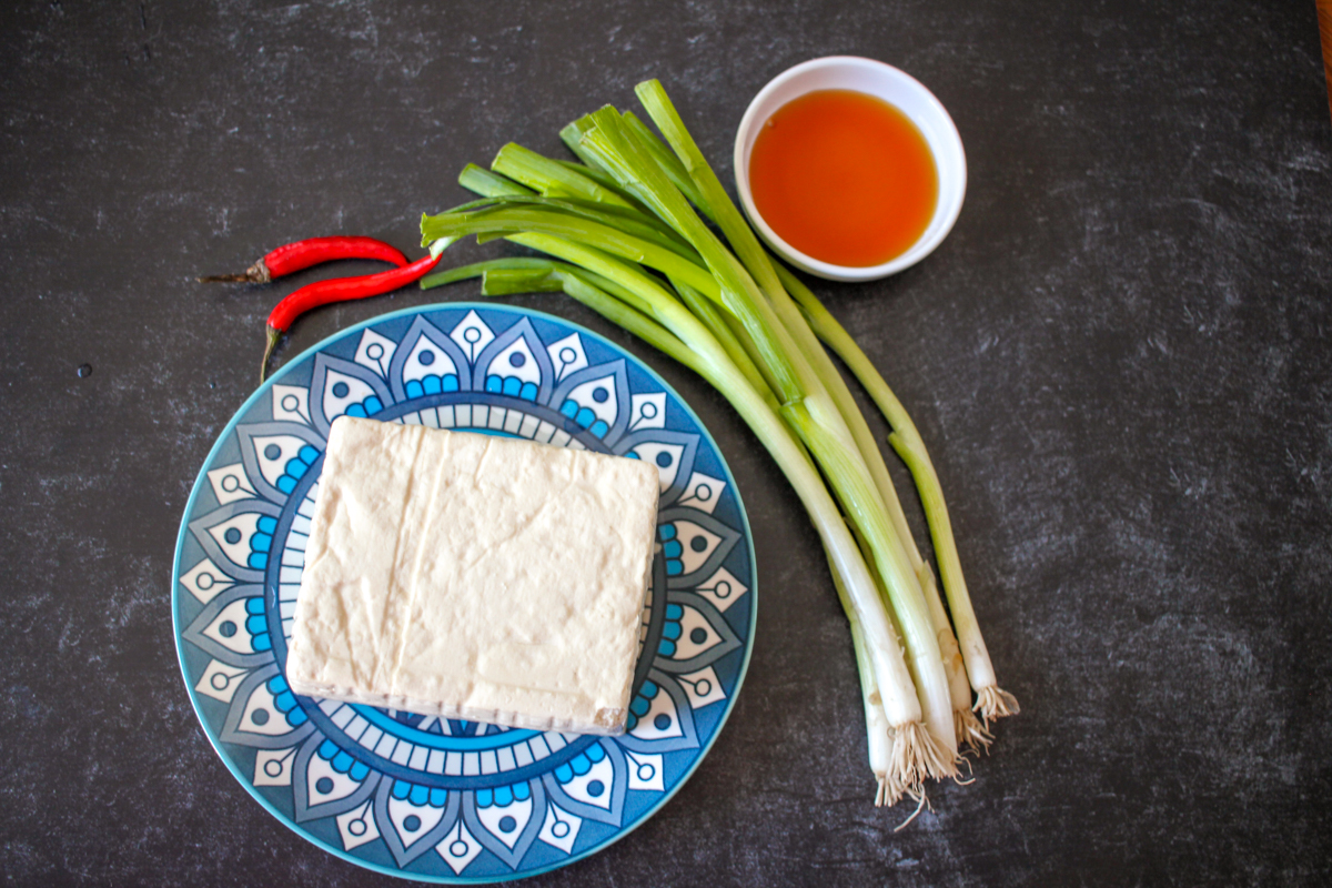 A block of tofu, two chilis, fish sauce, and a bunch of scallions arranged on a black background